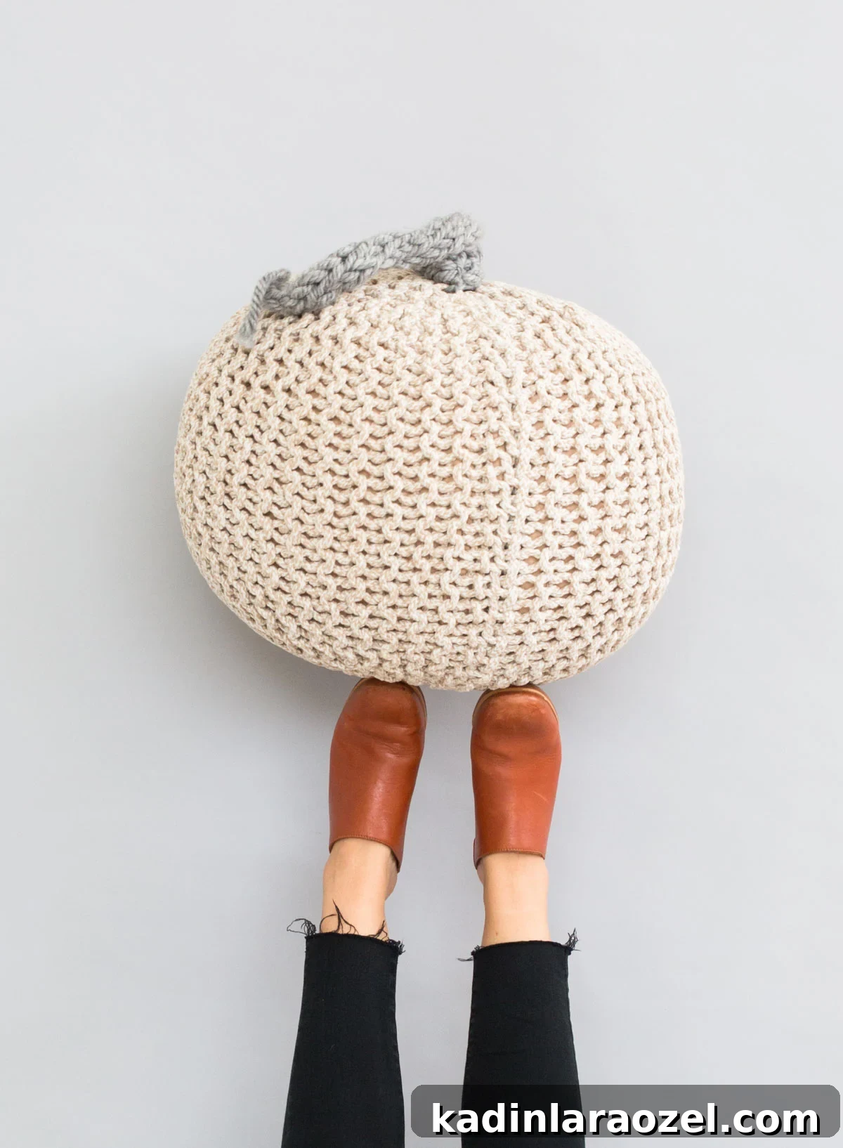 Neutral off white and grey giant knit pumpkin for Halloween, held up with feet against the wall