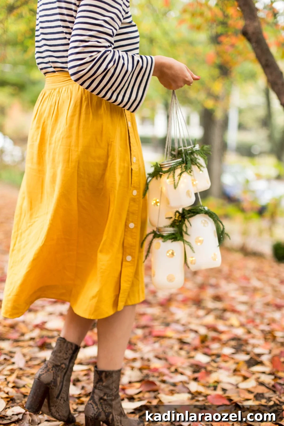 Woman walking outside in the fall leaves with boots on, carrying holiday hanging lanterns with fairy lights inside