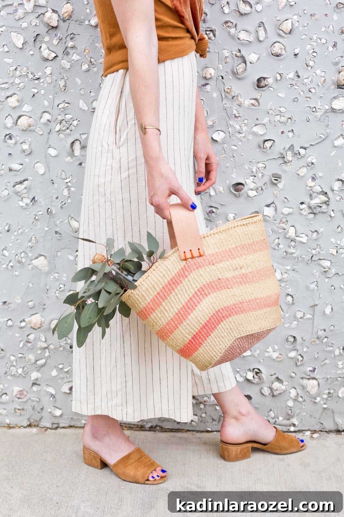 Woman wearing neutral clothing, carrying a striped basket bag that holds flowers and greenery