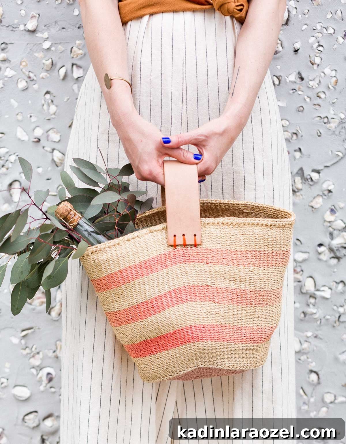 Woman carrying striped woven purse in front of her, that is holding a bottle of wine and floral greenery