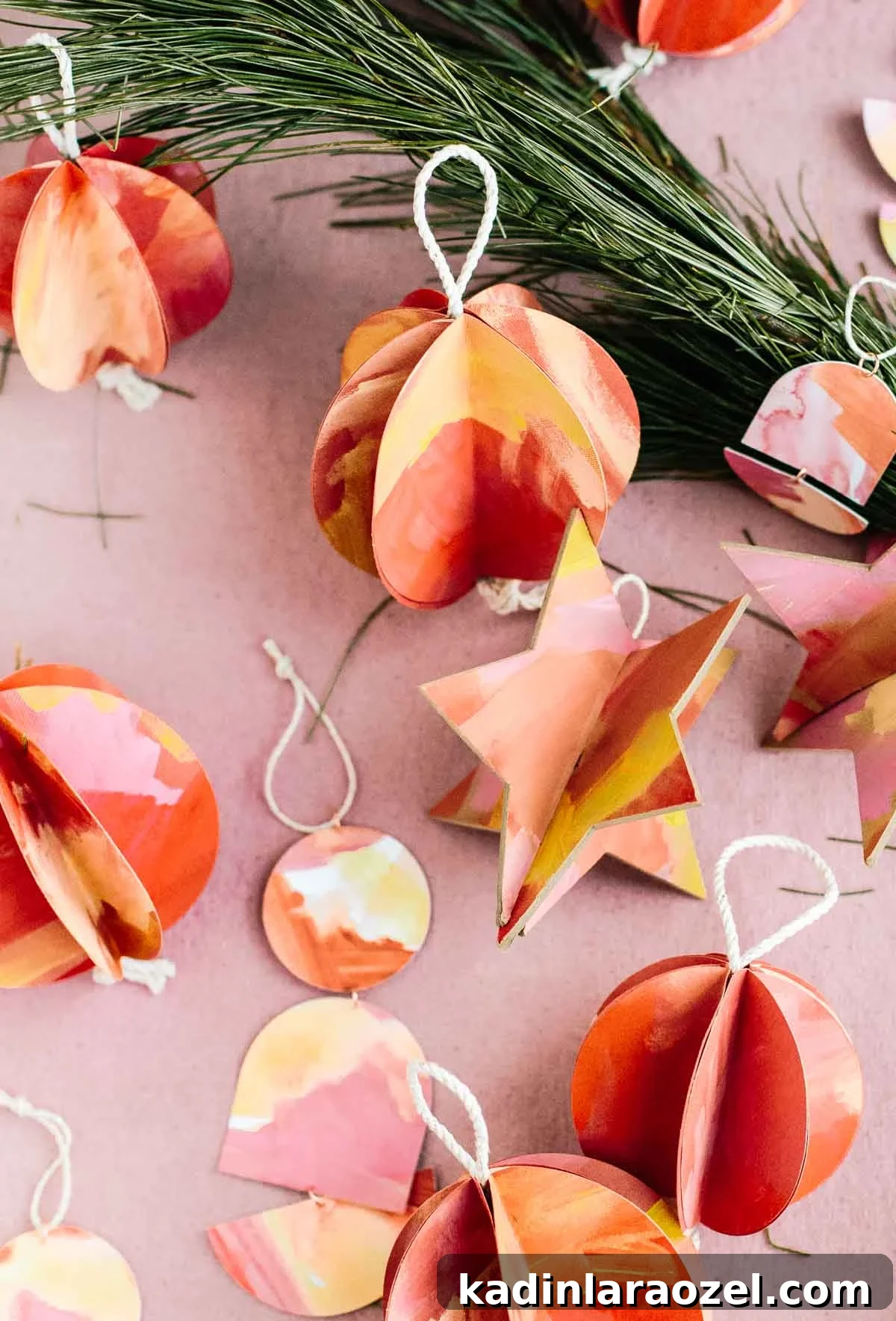 Christmas ornaments made of paper in various shapes laid out on a pink background with pine tree trimmings