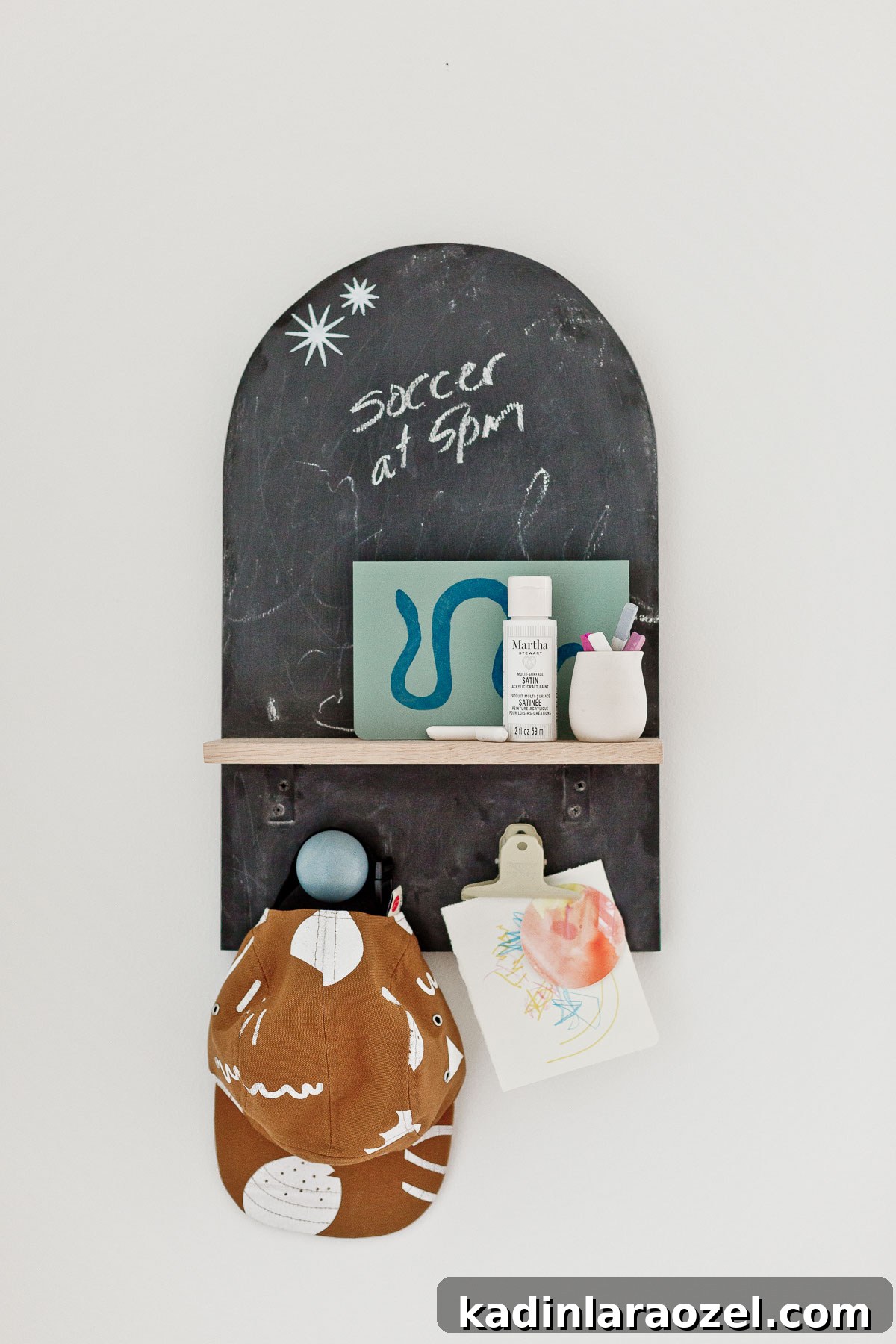 3 Stylish Back-to-School DIY Projects That Impress 11 Arch shaped chalkboard wall organizer hanging on white wall with kids hat, artwork, and art supplies on wooden shelf