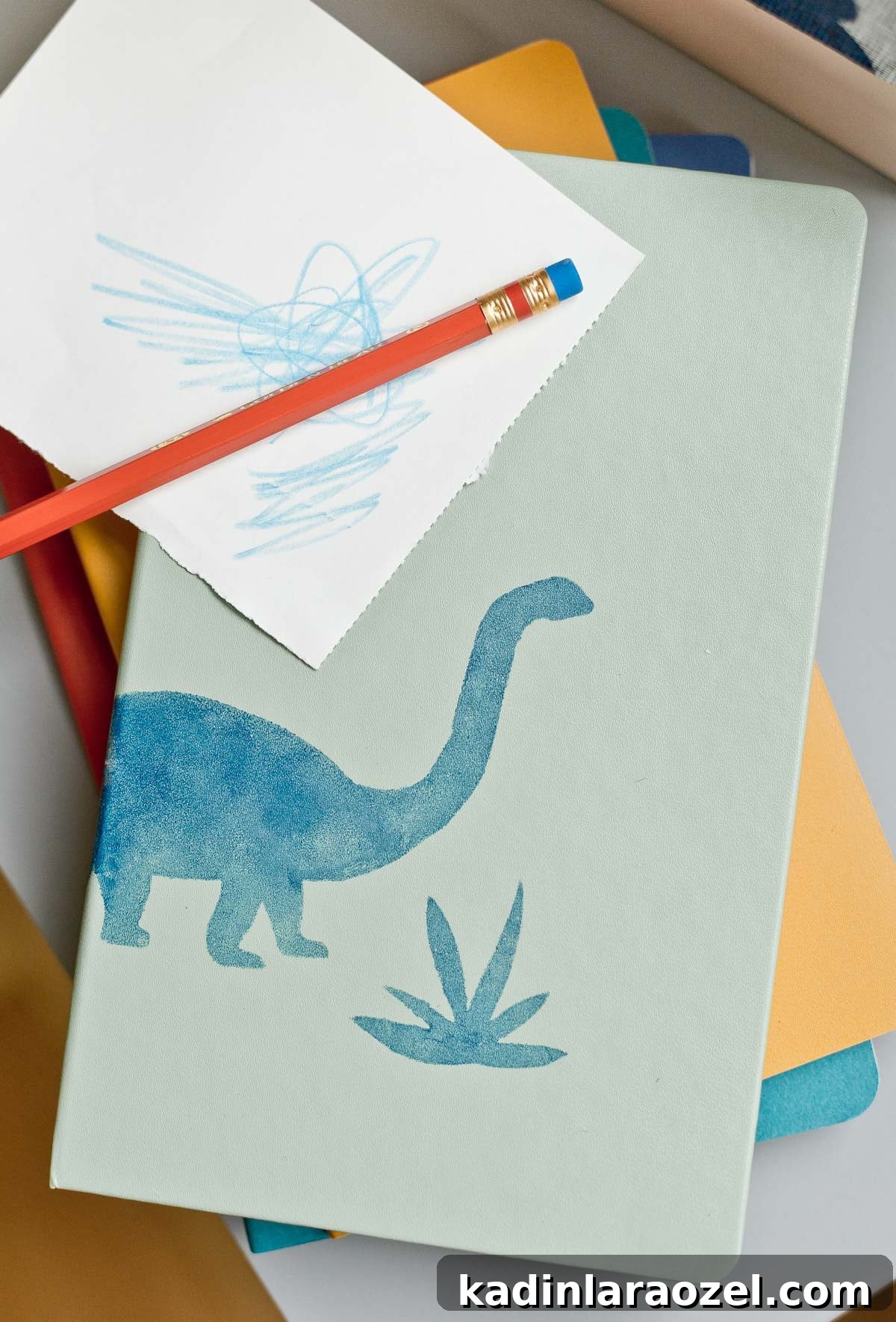 3 Stylish Back-to-School DIY Projects That Impress 9 Stack of colorful notebooks with dinosaur stencil design on it and a red pencil on top