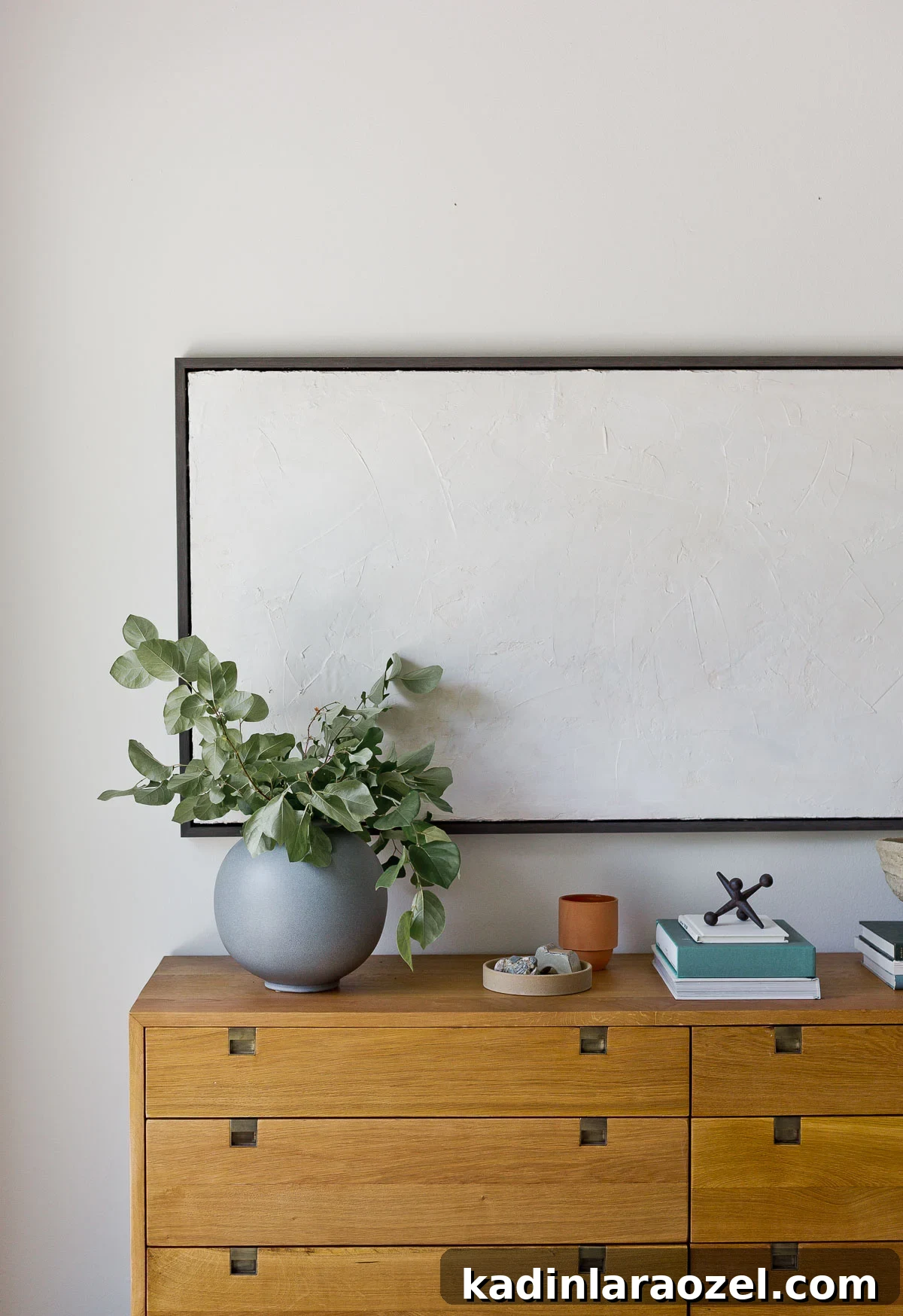 White living room with minimal artwork and a wood dresser that acts as a credenza, with books and a large grey vase.