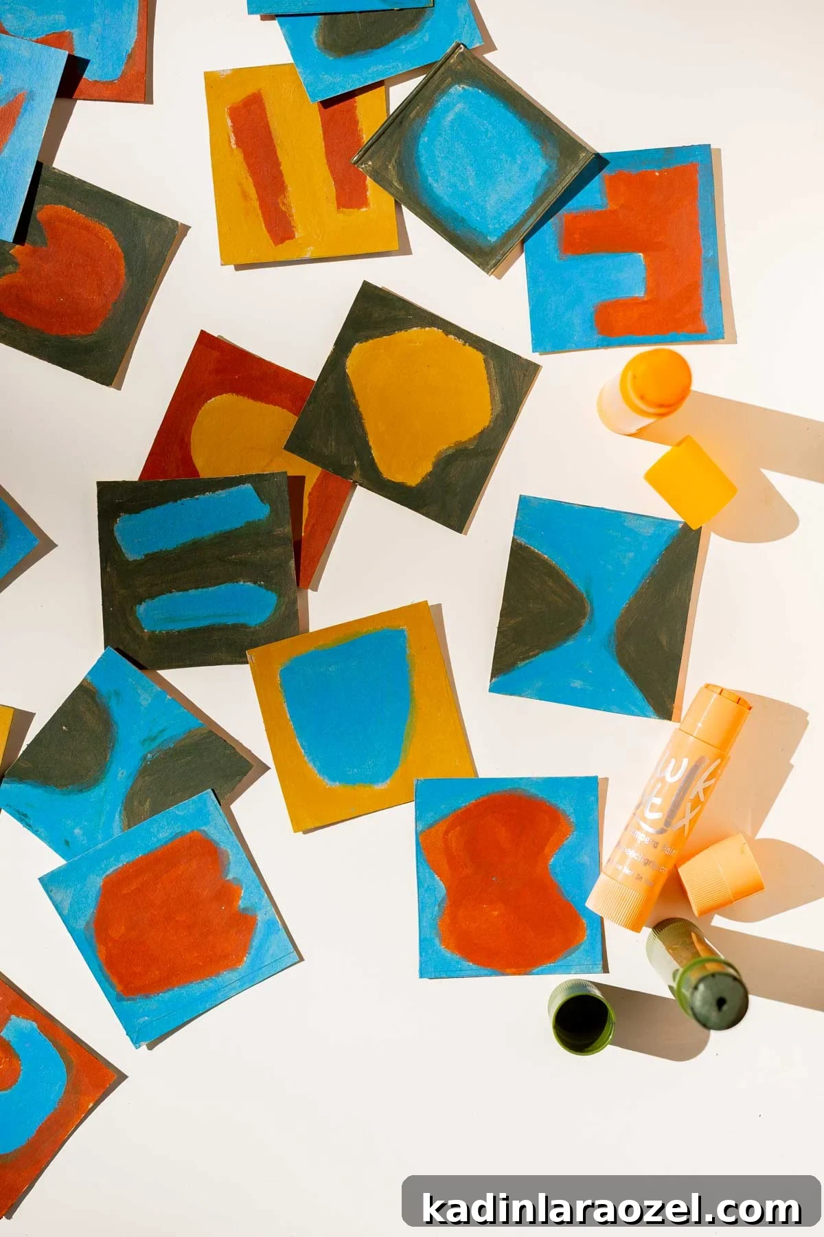 Small square paintings in abstract shapes laid out on a white table with art supplies